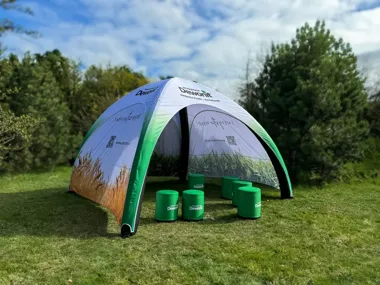 Nadmuchiwana kopuła reklamowa Dewont na trawie, otoczona zielonymi stołkami w parku.