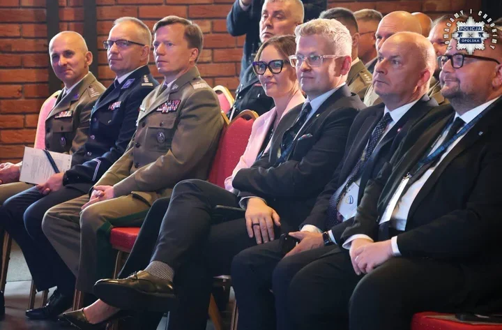 Grupa osób w mundurach i garniturach siedzi w rzędzie podczas oficjalnej uroczystości w sali z ceglanym tłem.