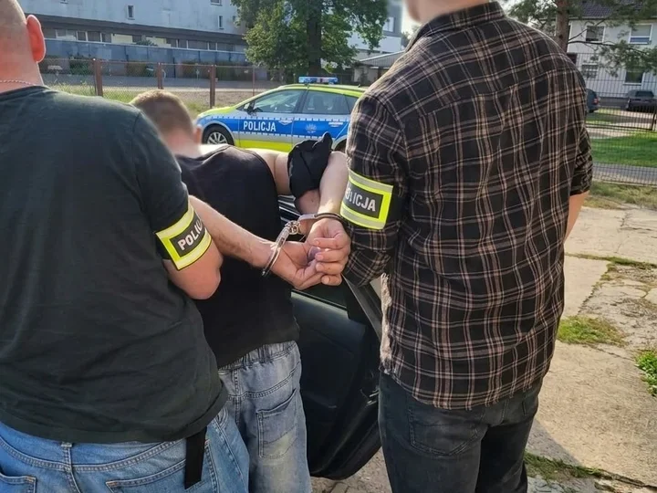 Funkcjonariusze policji zatrzymują mężczyznę w kajdankach przy policyjnym radiowozie na ulicy.