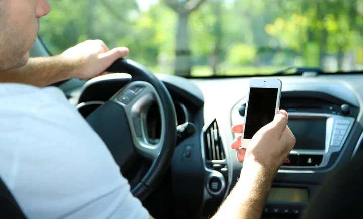 Kierowca w białej koszulce trzyma jedną ręką kierownicę, a w drugiej smartfon z czarnym ekranem, jadąc samochodem osobowym w słoneczny dzień.