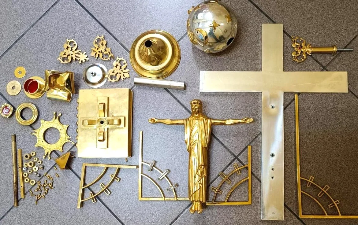 Rozłożone na szarym kafelkowanym podłożu złocone elementy krzyża procesyjnego z figurą Chrystusa, kulą zwieńczenia i detalami montażowymi.