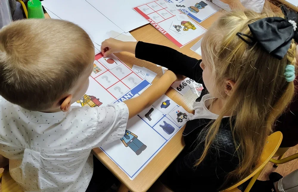 Dwoje dzieci siedzi przy drewnianym stoliku i układa elementy edukacyjnej gry planszowej z obrazkami związanymi z policją.