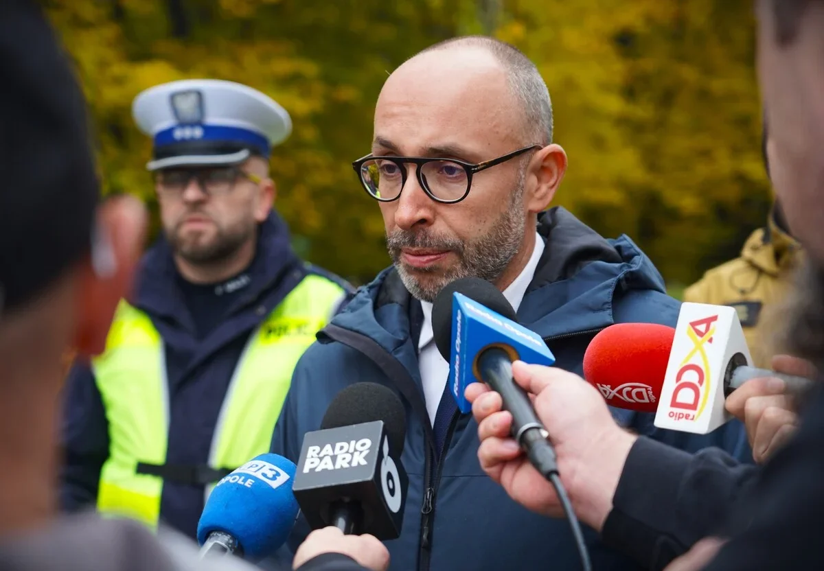 Mężczyzna w okularach i granatowej kurtce udziela wywiadu dziennikarzom z mikrofonami Radia Park, Radia Opole i innych mediów, w tle stoi policjant w kamizelce