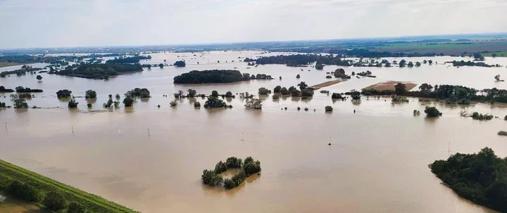 Rozległa powódź zalewa płaskie, rolnicze tereny, wystające z brązowej wody drzewa i niewielkie wyspy ziemi tworzą mozaikę zalanej doliny.
