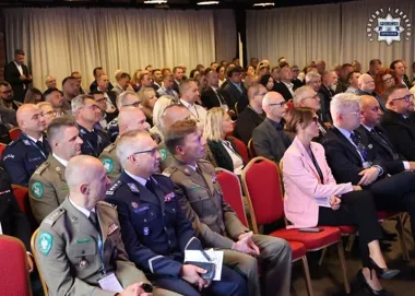 Uczestnicy oficjalnej uroczystości, w tym funkcjonariusze policji i służb mundurowych, siedzą na czerwonych krzesłach w sali konferencyjnej.