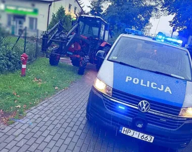 Policyjny radiowóz z włączonymi światłami stoi na brukowanej drodze obok ciągnika rolniczego w rejonie zabudowy mieszkaniowej.