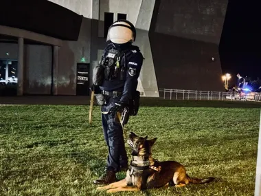 Funkcjonariusz policji w kasku z psem służbowym leżącym na trawie przed nowoczesnym budynkiem nocą.