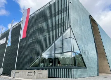 Nowoczesny budynek z charakterystyczną szklaną fasadą, z flagami przed wejściem oznaczonym napisem „Wejście/Entrance”.