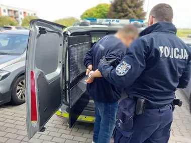 Funkcjonariusz policji w mundurze prowadzi zatrzymanego mężczyznę w kajdankach do tyłu radiowozu z otwartymi drzwiami, na parkingu przed budynkami mieszkalnymi.