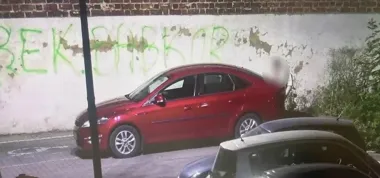 Czerwony samochód osobowy zaparkowany przy betonowej ścianie z zielonym graffiti, obok którego stoi rozmyta postać człowieka.