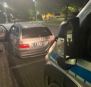 Policyjny radiowóz stoi przy srebrnym kombi BMW zaparkowanym na chodniku nocą na ulicy miasta.