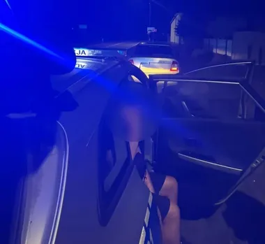 Nocna akcja policji na drodze: funkcjonariusz klęczy przy otwartych drzwiach samochodu osobowego, w tle radiowóz z włączonymi światłami.