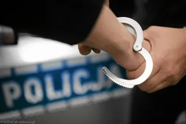 Funkcjonariusz policji trzyma kajdanki na tle oznakowanego radiowozu.