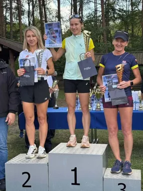 Trzy kobiety stoją na podium sportowym, trzymając trofea i dyplomy, podczas uroczystego wręczania nagród na zewnątrz w lesie.