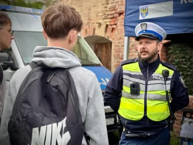 Policjant w kamizelce odblaskowej rozmawia z dwoma nastolatkami pod niebieskim namiotem z napisem „Marszałek Województwa Opolskiego”.