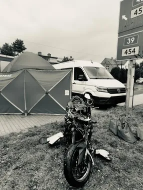 Uszkodzony motocykl leży na trawniku przy namiocie strażackim i białym furgonie, obok drogowskazu wskazującego kierunki dróg.