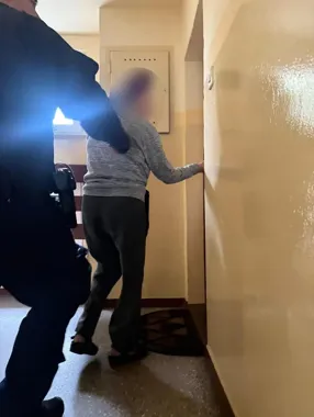 Funkcjonariusz w mundurze i osoba w szarej bluzie stoją przed drzwiami w klatce schodowej.