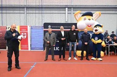 Funkcjonariusz policji przemawia do mikrofonu podczas uroczystego otwarcia sportowej imprezy w hali, obok stoją oficjele, policjanci i policyjne maskotki.