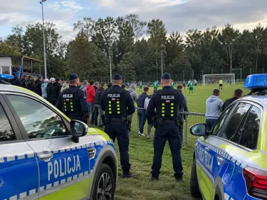Trzech policjantów w mundurach stoi między radiowozami i obserwuje mecz piłkarski na otwartym stadionie, za ogrodzeniem zgromadzeni kibicy.