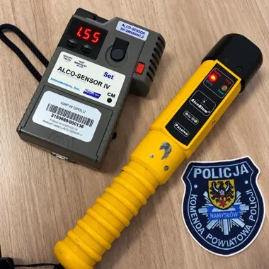 Sprzęt do badania trzeźwości: alkomat Alco-Sensor IV, tester AlcoBlow i naszywka policji z Namysłowa leżą na drewnianym blacie.