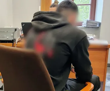 Mężczyzna w czarnej bluzie z kapturem siedzi plecami do obiektywu przy biurku w pomieszczeniu biurowym, naprzeciwko kobiety przy laptopie.
