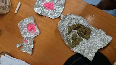 Na drewnianym stole leżą trzy kawałki folii aluminiowej z różowym proszkiem oraz suszonymi kwiatami konopi indyjskich, obok widoczna jest biała papierosowa skrę