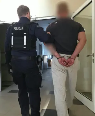 Policjant w mundurze z napisem POLICJA prowadzi zatrzymanego mężczyznę w kajdankach przez korytarz budynku.