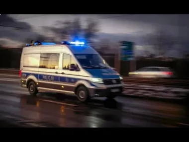 Policyjny radiowóz marki Volkswagen z napisem POLICJA jedzie z włączonymi niebieskimi światłami błyskowymi po mokrej drodze o zmierzchu.