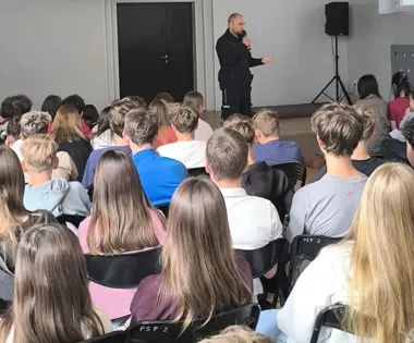 Mężczyzna w czarnym dresie przemawia do grupy nastolatków siedzących na krzesłach w sali z ciemnymi drzwiami w tle.