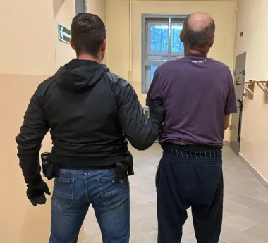 Funkcjonariusz w czarnej kurtce prowadzi mężczyznę w fioletowej koszulce korytarzem budynku.