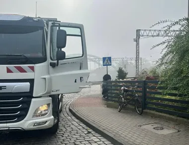 Biała ciężarówka DAF CF z otwartymi drzwiami stoi na brukowanej drodze obok ogrodzenia, przy którym oparto rower, w tle widoczny znak pieszy i mglista infrastru