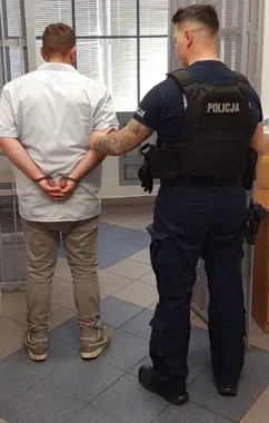 Funkcjonariusz policji w kamizelce z napisem „POLICJA” trzyma zatrzymanego mężczyznę w kajdankach w pomieszczeniu z kratami na oknach.