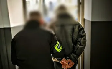 Funkcjonariusz policji w czarnej kurtce z żółtą opaską na ramieniu prowadzi zatrzymanego mężczyznę w kajdankach korytarzem budynku.