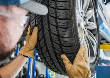 Mechanik w rękawicach i okularach ochronnych sprawdza stan bieżnika opony samochodu osobowego na podnośniku w warsztacie.
