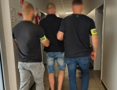 Funkcjonariusze policji prowadzą zatrzymanego mężczyznę w kajdankach korytarzem budynku.