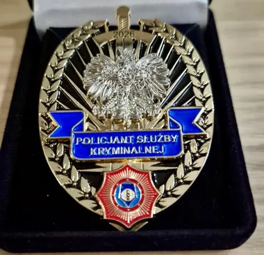 Odznaka „Policjant Służby Kryminalnej” z orłem, wstęgą i emblematem służby kryminalnej umieszczona w czarnym etui.