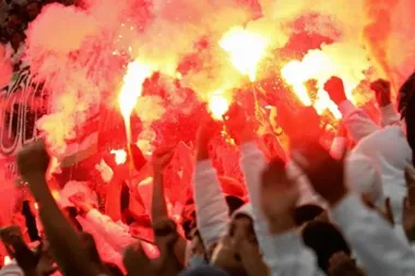 Tłum ludzi trzyma w górze zapalone race, wypełniając scenę intensywnym czerwonym światłem i dymem.