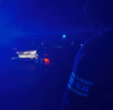 Policjant stoi przy samochodzie z włączonymi światłami podczas nocnej akcji drogowej.