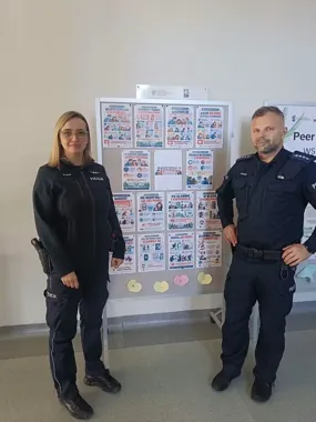 Dwoje policjantów w mundurach stoi po obu stronach tablicy z plakatami edukacyjnymi w pomieszczeniu.