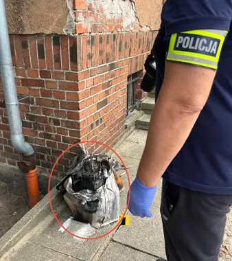 Policjant w niebieskich rękawiczkach stoi przy spalonym worku z niezidentyfikowanym przedmiotem na chodniku obok ceglanego budynku.