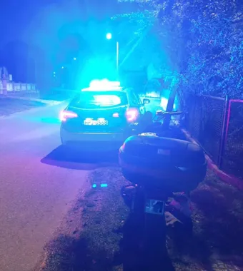 Policyjny radiowóz z włączonymi niebieskimi światłami stoi nocą na poboczu drogi obok zaparkowanego skutera.