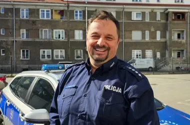 Uśmiechnięty policjant w granatowym mundurze stoi przed policyjnym radiowozem na tle wielorodzinnego budynku.