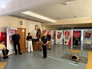 W sali szkolnej odbywa się spotkanie edukacyjne z udziałem policjantów i strażaków, obok leży fantom do nauki pierwszej pomocy.