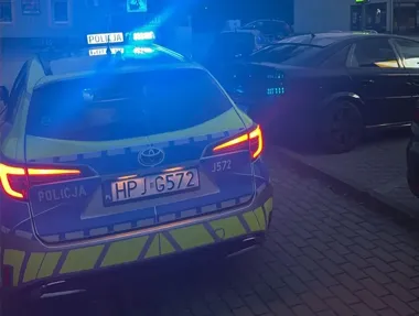 Policyjny radiowóz marki Toyota z włączonymi niebieskimi światłami stoi na brukowanej ulicy obok zaparkowanych samochodów nocą.