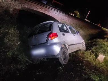 Srebrny samochód osobowy z uszkodzoną tylną szybą stoi na skarpie przy torach kolejowych nocą.