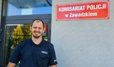 Uśmiechnięty policjant w granatowej koszulce polo z napisem „POLICJA” stoi przed budynkiem komisariatu policji w Zawadzkiem.
