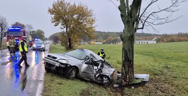 Strażacy i policjanci przy rozbitym srebrnym samochodzie osobowym, który uderzył w drzewo przy wiejskiej drodze podczas deszczu.
