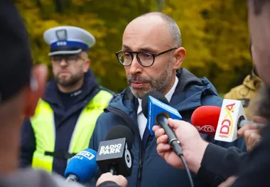 Mężczyzna w okularach i granatowej kurtce udziela wywiadu dziennikarzom z mikrofonami Radia Park, Radia Opole i innych mediów, w tle stoi policjant w kamizelce