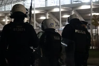 Trzech policjantów w kaskach i umundurowaniu stoi tyłem przed oświetloną trybuną stadionu nocą.