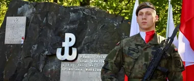 Żołnierz Wojska Polskiego w kamuflażu stoi z bronią przed pomnikiem Armii Krajowej i Powstania Warszawskiego podczas uroczystości z udziałem flag.